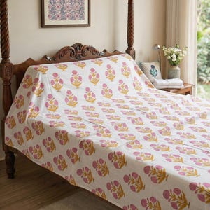 May include: A white bedspread featuring a repeating floral pattern of pink blossoms with yellow stems and leaves. The fabric is draped over a dark wooden bed frame, covering the mattress and extending down the sides with a consistent botanical print.