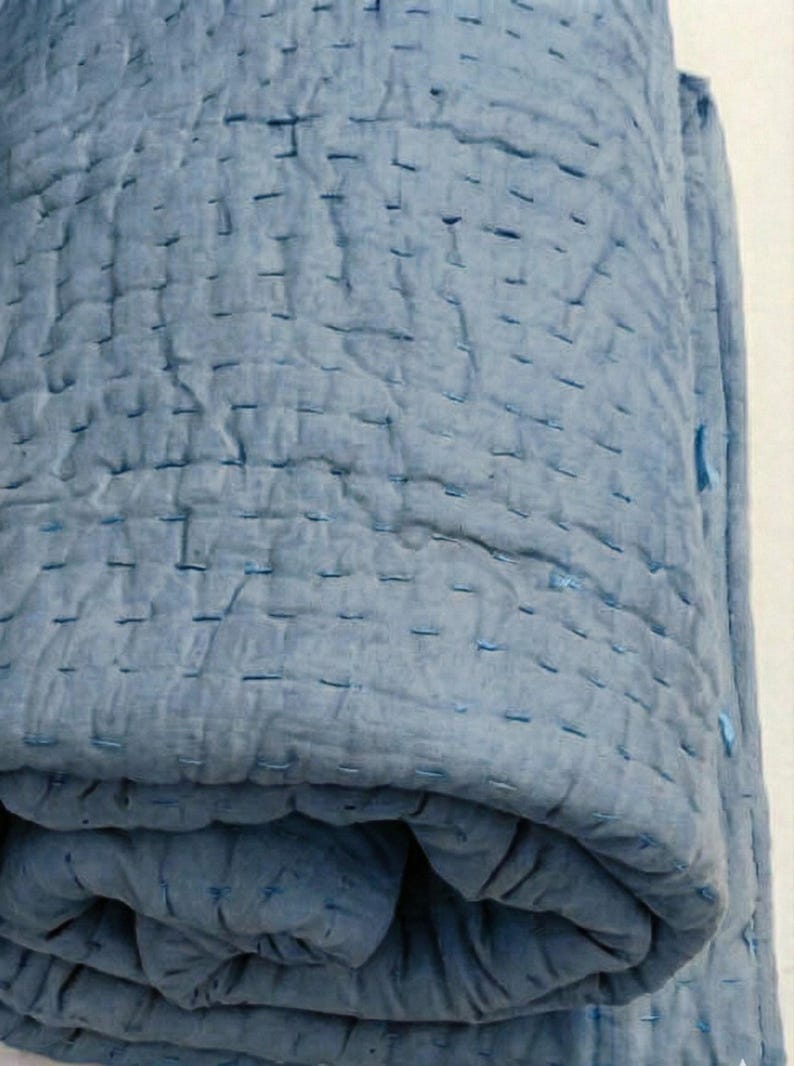 May include: A rolled-up, light blue quilt with visible stitching. The quilt appears to be made of a soft, textured fabric. The stitching is a darker shade of blue, creating a subtle contrast. The overall aesthetic is simple and cozy.