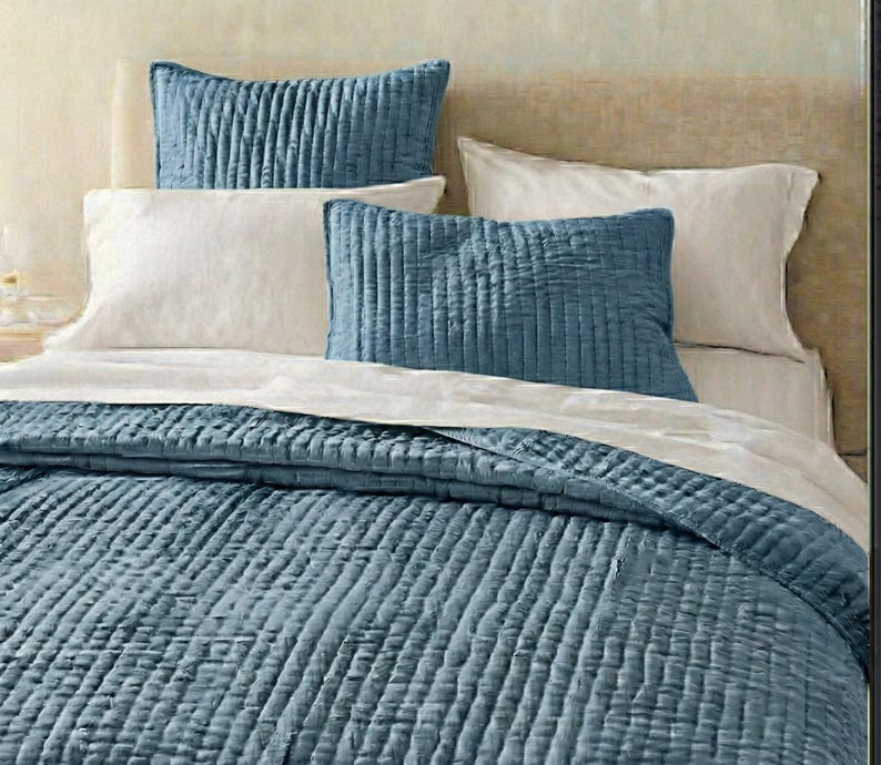 May include: A bed with a blue quilted comforter and matching pillows. The pillows are rectangular and have a vertical ribbed texture. White pillowcases and a white sheet are also visible. The bed is against a neutral-toned headboard.