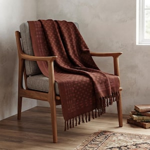 May include: A deep rust-colored throw blanket with a subtle repeating geometric dot pattern and fringed edges, draped over a wooden armchair with gray upholstered cushions. The blanket features a distinct border design along the sides.