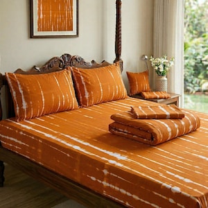 May include: A bedding set featuring a burnt orange colour with white tie-dye style vertical stripes. The set includes a fitted sheet, two rectangular pillowcases, a square throw pillow, and a folded comforter, all matching in pattern and colour.