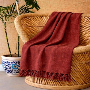 May include: A terracotta-coloured woven throw blanket with a fringed edge, draped over a wicker chair. A potted plant with green leaves is to the left. The blanket has a textured look and is made of a soft material.