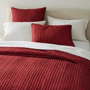 Könnte beinhalten: Ein Bett mit einer rot gesteppten Bettdecke und passenden Kissenbezügen. Weiße Kissenbezüge und ein weißes Laken sind ebenfalls zu sehen. Das Kopfteil ist in einem neutralen Beige gehalten. Die Bettwäsche hat ein strukturiertes Rippenmuster.
