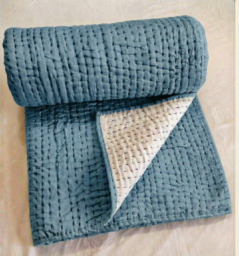May include: A rolled-up, blue quilted blanket with a white interior. The blanket is made of a soft, textured fabric and features a grid-like stitching pattern. The corner is folded back, revealing the white interior.