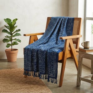 May include: A rectangular throw blanket featuring a deep blue fabric with a white intricate botanical pattern and a decorative border. The blanket has a fringed edge with knotted tassels hanging from the bottom, draped over a wooden chair.