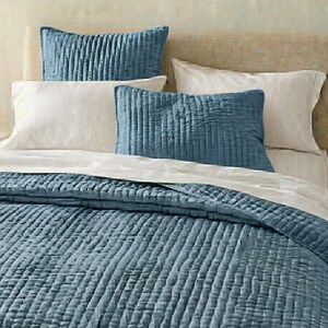 May include: A bed with a blue quilted comforter and matching pillows. The pillows are rectangular and have a vertical ribbed texture. White pillowcases and a white sheet are also visible. The bed is against a neutral-toned headboard.