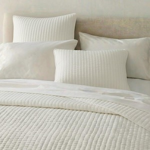 May include: A bed dressed in white bedding, featuring a quilted comforter, a flat sheet, and multiple pillows. The pillows have both plain and textured designs. The overall style is clean and minimalist, emphasising comfort and simplicity.
