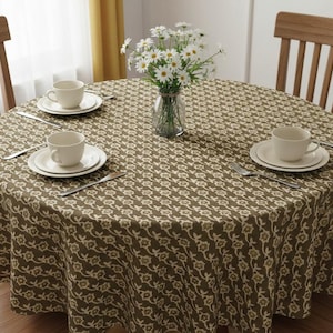 Puede incluir: Un mantel redondo con un estampado floral repetido en tonos marrón y crema cubre una mesa de madera. Tazas de té blancas, platillos y cubiertos están puestos para cuatro personas. Un jarrón con flores blancas está en el centro.