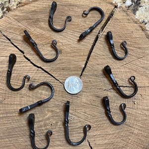 May include: Eight black metal hooks with a twisted design, arranged in a circle on a wooden surface. A US quarter is included for scale.