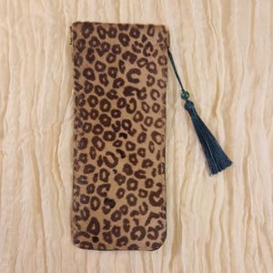Puede incluir: Un estuche rectangular para gafas con un patrón de estampado de leopardo en tonos beige y marrón. El estuche presenta un cordón fino sujeto en la esquina superior, que incluye una pequeña cuenta verde y una borla de tela azul verdoso oscuro.