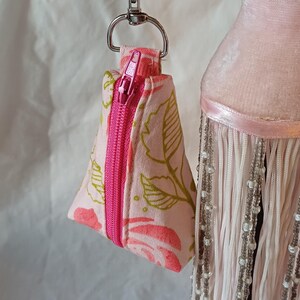 May include: A small, triangular pouch with a pink zip and a silver clasp. The pouch is made of pink fabric with a floral pattern in shades of pink and green. The zip is bright pink.