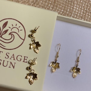Op de afbeelding: Goudkleurige blad oorbellen gepresenteerd in een crèmekleurige doos met de tekst "SALT SAGE & SUN". De oorbellen liggen op een witte ondergrond, met een jute achtergrond.