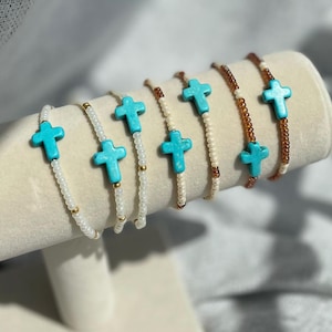 Op de afbeelding: Een display van armbanden met turquoise kruisjes. De armbanden hebben witte, gouden en bruine kralen. Ze worden getoond op een crèmekleurige, cilindrische standaard tegen een zachte, neutrale achtergrond.