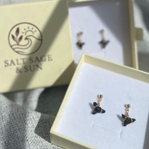 Puede incluir: Pendientes exhibidos en cajas crema abiertas sobre una superficie gris texturizada. Los pendientes presentan un pequeño dije de color oscuro suspendido de un poste dorado. Las cajas tienen el logotipo "SALT SAGE & SUN" impreso.