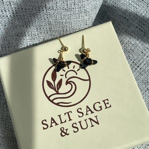 May include: Gold-colored earrings with black, star-shaped stones. The earrings are displayed on a cream-colored box with the text "SALT SAGE & SUN" and a logo featuring a wave and sun design.