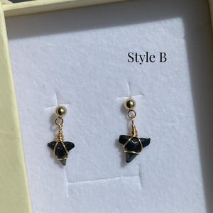 May include: A pair of gold-colored earrings with black, triangular pendants, possibly shark teeth. The earrings are displayed in a light-colored box, with the text "Style B" visible.