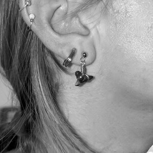 May include: Close-up of an ear adorned with multiple earrings. A small, silver shark tooth earring hangs from the lobe. Other earrings include a small hoop and a stud. The image is in black and white.