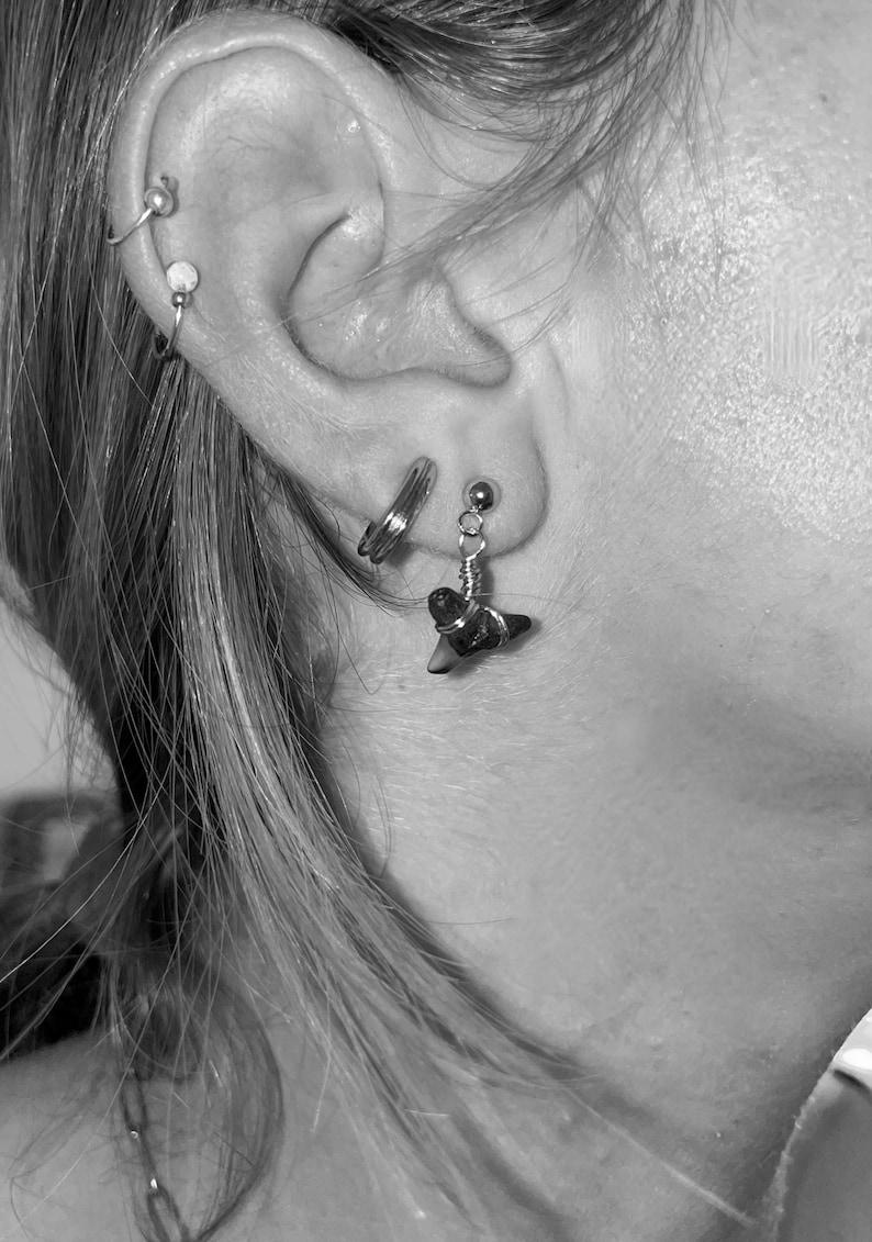 May include: Close-up of an ear adorned with multiple earrings. A small, silver shark tooth earring hangs from the lobe. Other earrings include a small hoop and a stud. The image is in black and white.