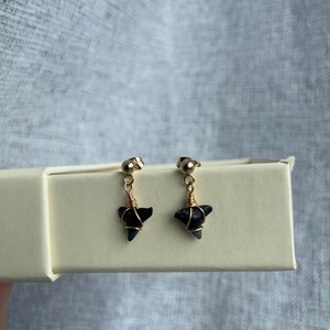 May include: A pair of gold-colored stud earrings with small, dark gray, shark tooth-shaped pendants. The pendants are wrapped in gold wire and hang from the studs. The earrings are displayed on a light-colored box.