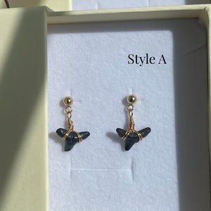 May include: A pair of earrings featuring small, dark gray shark teeth. The teeth are wrapped in gold-colored wire and suspended from gold-colored ball studs. The text "Style A" is visible in the background.