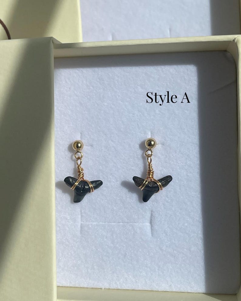 May include: A pair of earrings featuring small, dark gray shark teeth. The teeth are wrapped in gold-colored wire and suspended from gold-colored ball studs. The text "Style A" is visible in the background.
