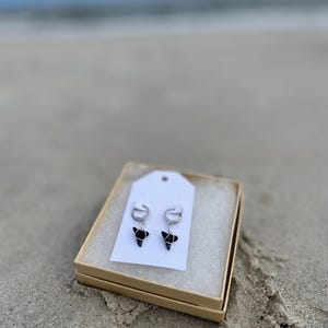 Könnte beinhalten: Ein Paar schwarze Haifischzahn-Ohrringe mit silbernen Creolen, präsentiert in einer kleinen, offenen Pappschachtel mit weißer Polsterung. Die Schachtel befindet sich an einem Sandstrand mit dem Meer im Hintergrund.