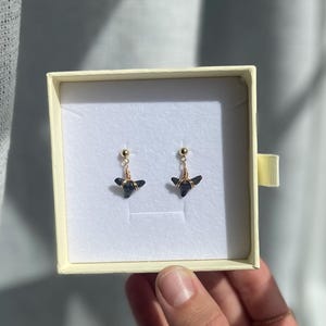 May include: A pair of black gemstone earrings with gold-colored wire and post detailing. The earrings are displayed inside a cream-colored jewelry box with a gold-colored ribbon. The earrings have a unique, geometric shape.
