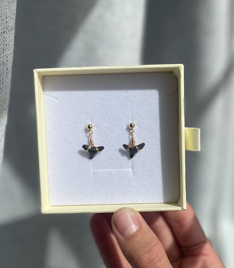 May include: A pair of black gemstone earrings with gold-colored wire and post detailing. The earrings are displayed inside a cream-colored jewelry box with a gold-colored ribbon. The earrings have a unique, geometric shape.