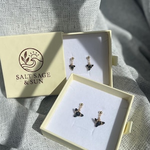 May include: Earrings displayed in open cream-colored boxes. The earrings have a gold-colored post and a black, geometric design. The boxes are placed on a textured, gray surface. One box has the text "SALT SAGE & SUN" with a logo.
