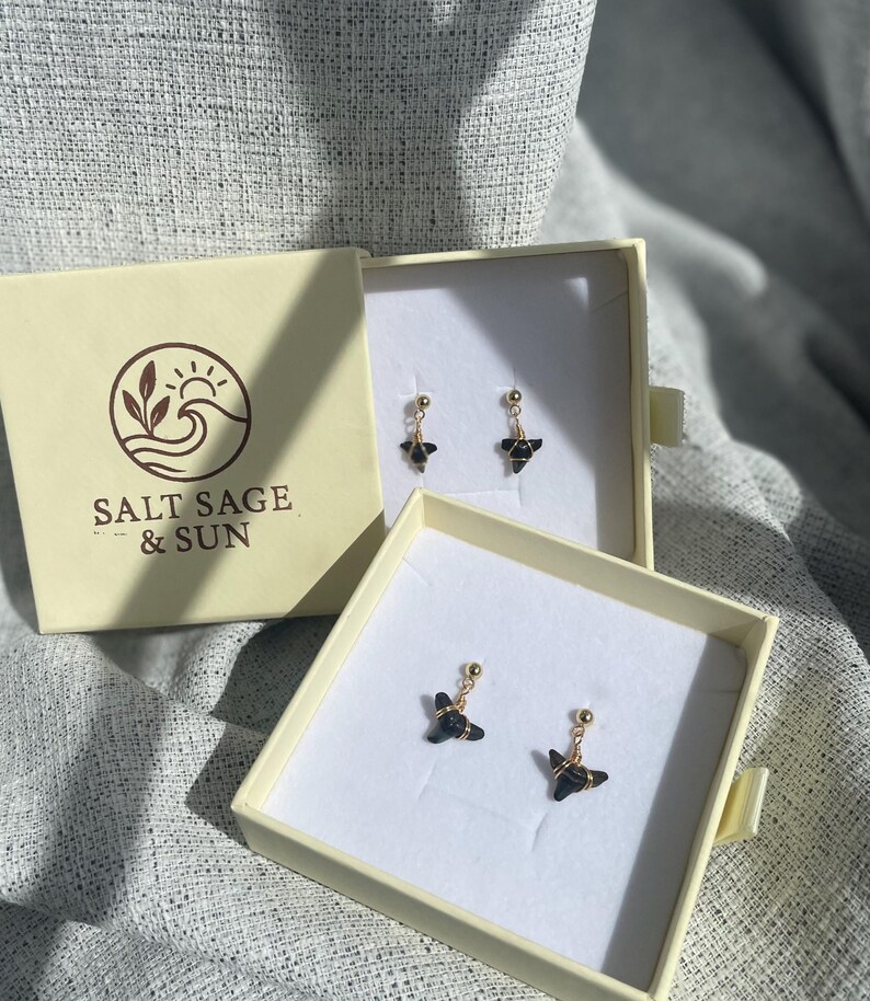 May include: Earrings displayed in open cream-colored boxes. The earrings have a gold-colored post and a black, geometric design. The boxes are placed on a textured, gray surface. One box has the text "SALT SAGE & SUN" with a logo.