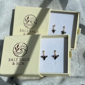 May include: Two pairs of earrings in open cream-colored boxes. The earrings have gold-colored posts and wires, with black, triangular-shaped stones. The boxes are labeled "SALT SAGE & SUN" and "SALT SAGE & OLIVE" with a logo of a wave, sun, and leaves.