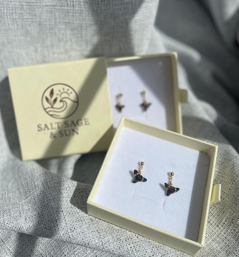 May include: Earrings displayed in open cream-colored boxes on a textured gray surface. The earrings feature a small, dark-colored charm suspended from a gold-colored post. The boxes have the logo "SALT SAGE & SUN" printed on them.