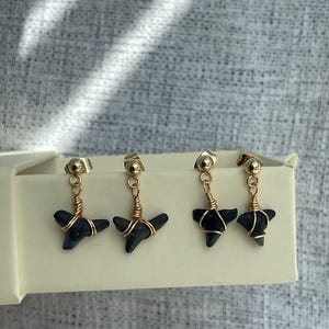 May include: Four gold-colored stud earrings with black, shark tooth-shaped pendants. The pendants are wrapped in copper-colored wire. The earrings are displayed in a cream-colored box against a gray background.