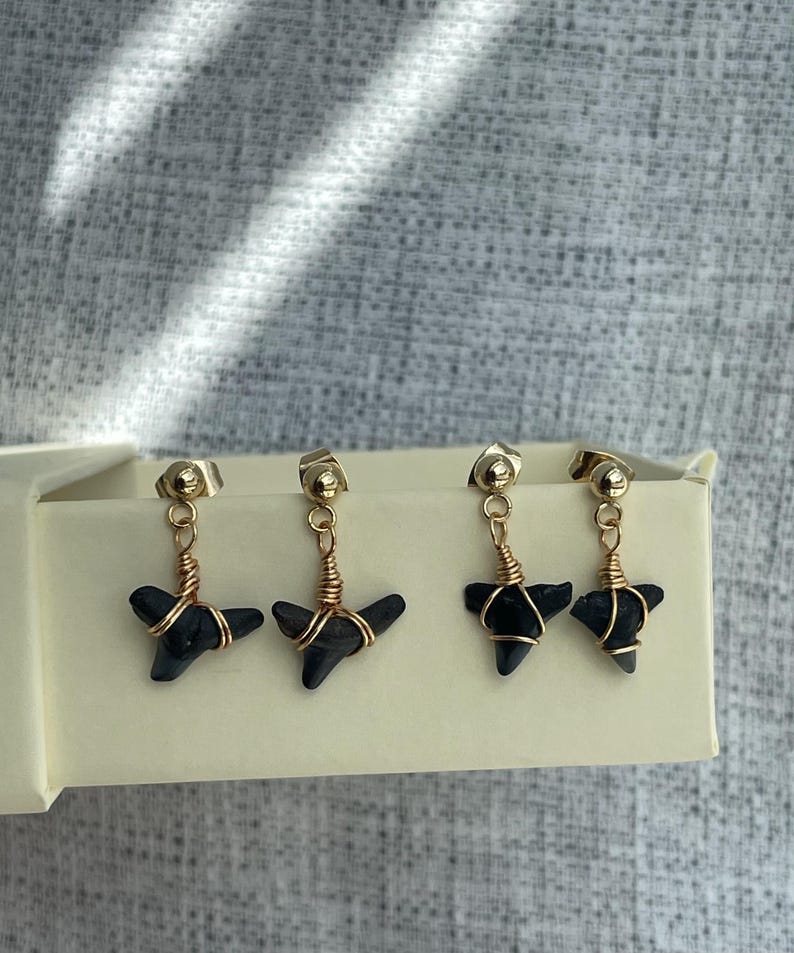May include: Four gold-colored stud earrings with black, shark tooth-shaped pendants. The pendants are wrapped in copper-colored wire. The earrings are displayed in a cream-colored box against a gray background.