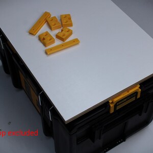 May include: A black and yellow plastic tool storage box with a white rectangular worktop surface. On top of the worktop are several small yellow plastic components with pre-drilled holes. The text Worktop excluded is printed in red.