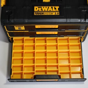 May include: Black and yellow DEWALT TOUGH SYSTEM 2.0 storage box with an open drawer revealing a grid of yellow compartments. The box has a black handle and the DEWALT logo in yellow. The open drawer is filled with yellow compartments.
