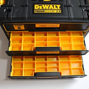 Puede incluir: Caja de almacenamiento de herramientas negra y amarilla DeWalt ToughSystem 2.0 con dos cajones abiertos. Los cajones están llenos de compartimentos amarillos. El logotipo de DeWalt y "TOUGHSYSTEM 2.0" están impresos en amarillo en la tapa negra.