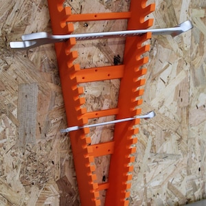 May include: A bright orange wall-mounted spanner organiser featuring a vertical, tapered design with multiple notched slots along both sides to hold various tools. Two silver metal spanners are positioned horizontally within the slots.