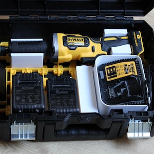 Puede incluir: Un maletín de herramientas DeWalt negro y amarillo, abierto para revelar una lámpara de trabajo, dos cargadores de batería y dos baterías. El kit está organizado con divisores blancos y amarillos. El logotipo de DeWalt es visible en las herramientas y los cargadores.