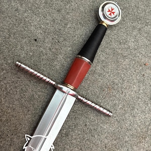 May include: A medieval-style sword with a silver blade and a red and black handle. The hilt features a cross-shaped design. The pommel has a white and silver design with a red cross. The text "Night Shade Gift" is visible on the blade.