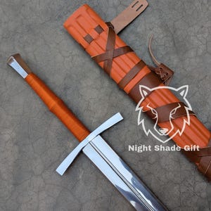May include: A medieval sword with a silver blade and an orange wrapped handle. The sword is accompanied by an orange sheath secured with brown leather straps. The image includes the text "Night Shade Gift".