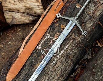 May include: A silver sword with a decorative hilt and crossguard, resting on a log. The sword has a long, polished blade and is accompanied by a brown leather sheath. The text "Night Shade Gift" is visible on the blade.