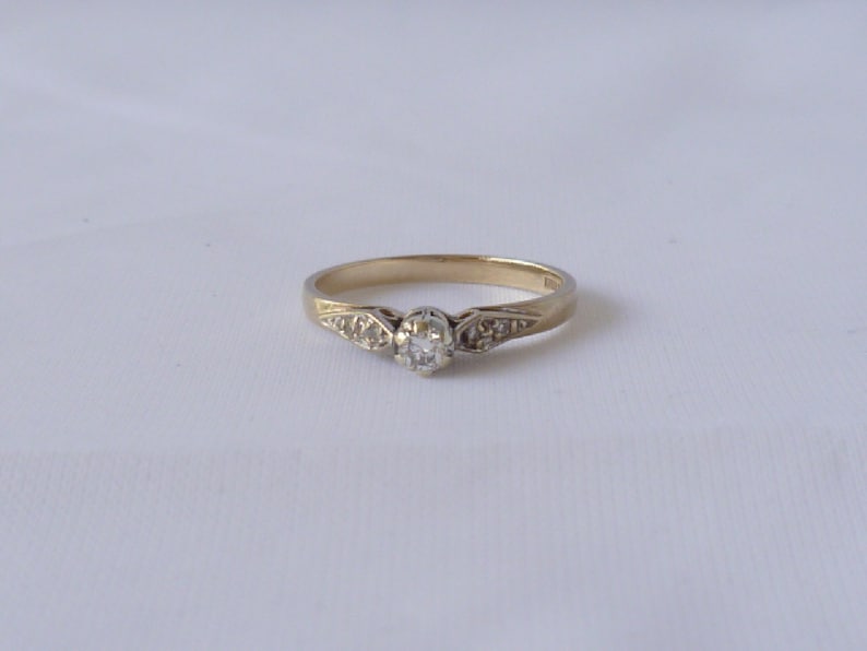 May include: A delicate gold ring with a single round diamond set in the center. The band is adorned with smaller diamonds on either side of the center stone.