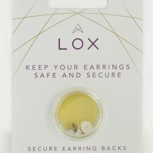 Puede incluir: Una tarjeta blanca con patrones geométricos dorados y el texto "A LOX KEEP YOUR EARRINGS SAFE AND SECURE" en morado. Se muestra un recipiente de plástico transparente con dos pares de cierres de pendientes de tono dorado.