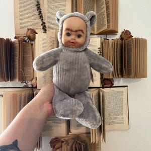 Könnte beinhalten: Eine graue Plüschpuppe mit kahlem Kopf, blauen Augen und einem kleinen Bart, die ein Kapuzen-Bärenkostüm trägt. Die Puppe wird vor einer Wand gehalten, die mit offenen Büchern und getrockneten Rosen dekoriert ist.