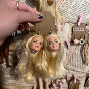 Könnte beinhalten: Ohrringe mit Puppenköpfen mit blondem Haar, blauen Augen und rosa Lippen. Sie sind an kleinen braunen Anhängern befestigt. Weitere Ohrringe mit Puppenteilen sind zu sehen, sowie Puppenbeine. Der Hintergrund zeigt Textseiten.