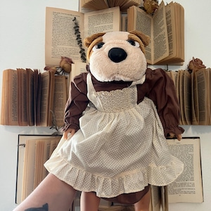 May include: A plush bulldog doll wearing a brown dress with a white apron featuring a polka dot pattern. The doll has a tan and brown head with a black nose. The doll is held in front of a wall decorated with open books.