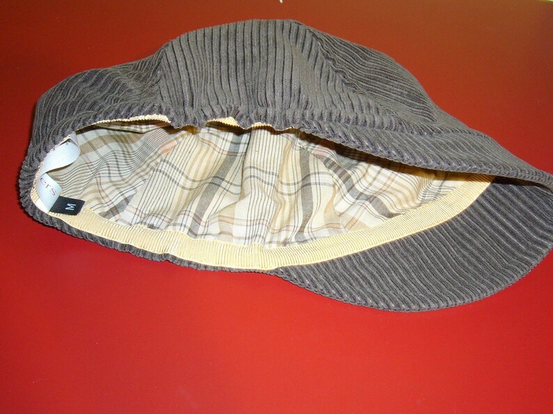 Puede incluir: Una gorra de pana marr&oacute;n con forro a cuadros y una banda el&aacute;stica amarilla. La gorra tiene una etiqueta de talla que dice "M".