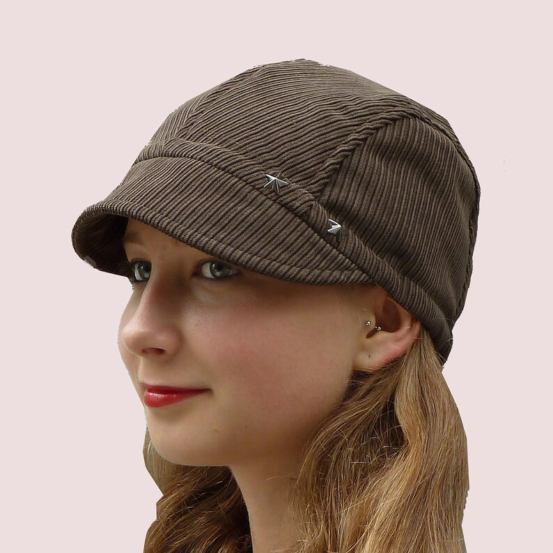 Puede incluir: Una gorra de newsboy de pana marr&oacute;n con una visera ligeramente curvada y dos adornos en forma de estrella plateada.