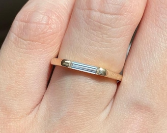 May include: A gold band ring featuring a single, horizontally set, clear baguette-cut diamond at the center. The ring has a smooth, polished finish and a slim profile, showcasing a clean, linear design that sits flush against the finger.
